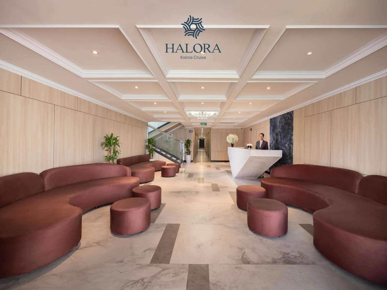 Sảnh lễ tân Halora Kairos với trần ốp gỗ dạng ô caro sang trọng, hai dãy sofa cong màu nâu đỏ bố trí đối xứng trên nền đá marble, dẫn lối tới quầy lễ tân đá trắng hình nêm ở cuối sảnh.
