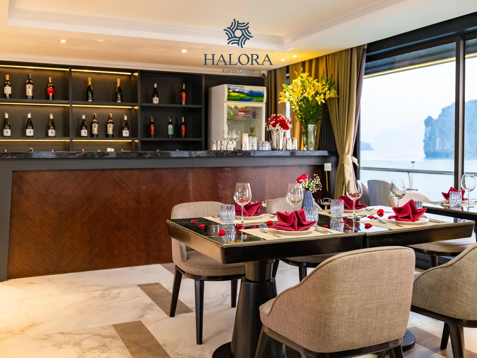Nhà hàng sang trọng với quầy bar rượu vang đèn vàng ấm áp phía sau, bàn tiệc bày khăn ăn đỏ gấp hình hoa cùng bình hoa ly vàng rực rỡ, bên khung cửa kính lớn phóng tầm mắt ra vịnh đá vôi.