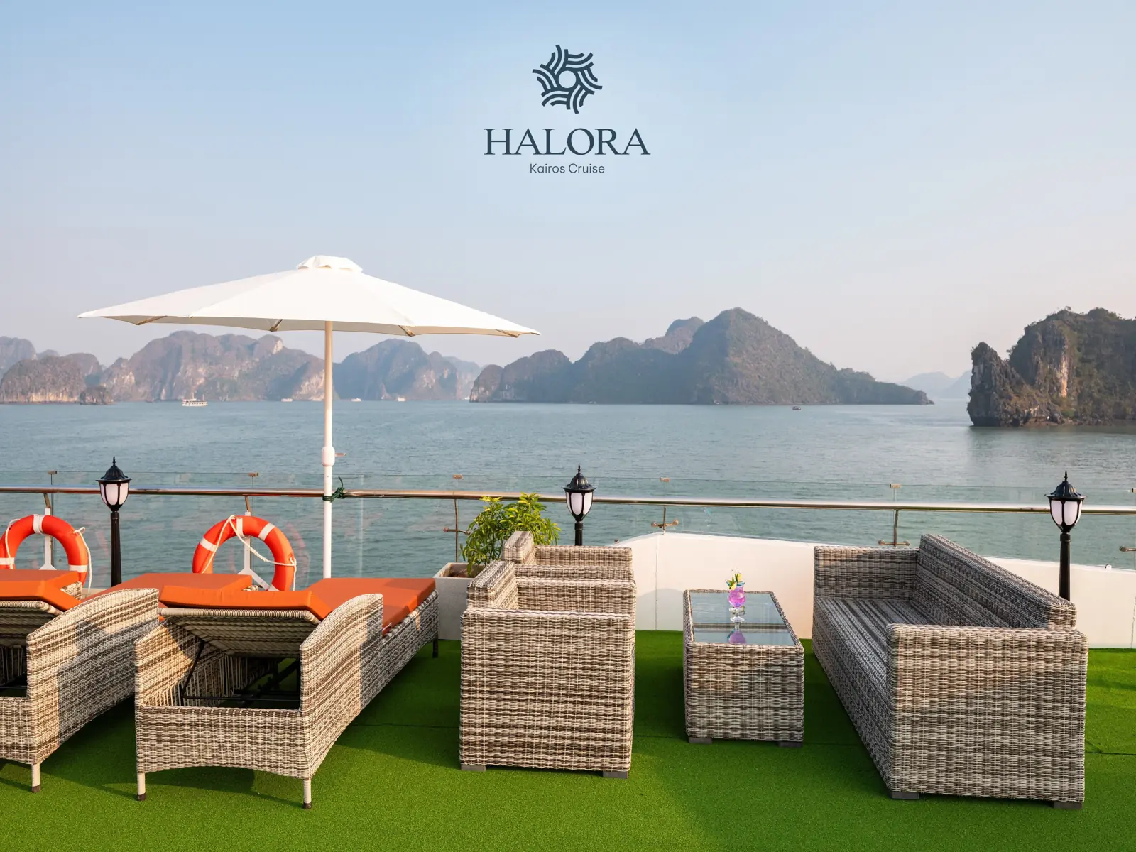 Góc thư giãn trên sundeck với ô dù trắng lớn, bộ sofa mây đệm cam cùng phao cứu sinh đỏ nổi bật trên nền cỏ nhân tạo xanh mướt, phóng tầm mắt ra vịnh với những dãy núi đá vôi hùng vĩ.