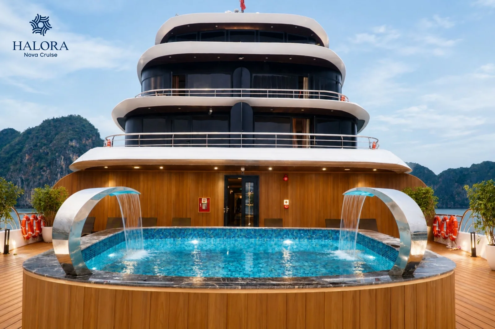 Hồ bơi vô cực hình oval nằm trên sun deck với hai vòi phun nước cong bằng thép không gỉ tạo thác đổ mềm mại, bao quanh là sàn gỗ teak và tầm nhìn ra núi đá vịnh Lan Hạ dưới nắng.